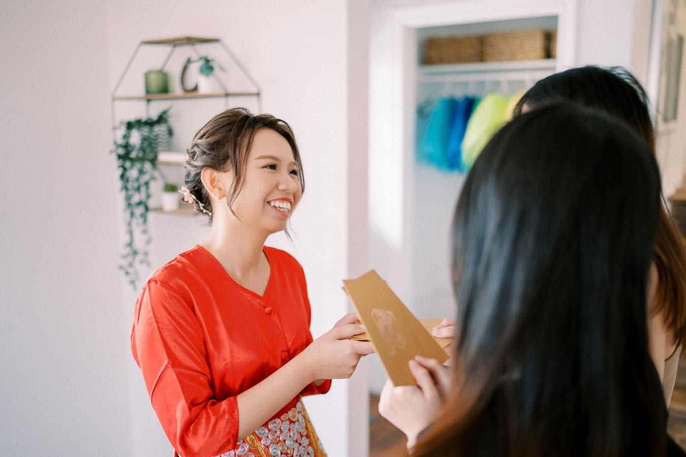 Smiling woman in a red dress exchanging a gift envelope with friends indoors, symbolizing celebration and friendship.