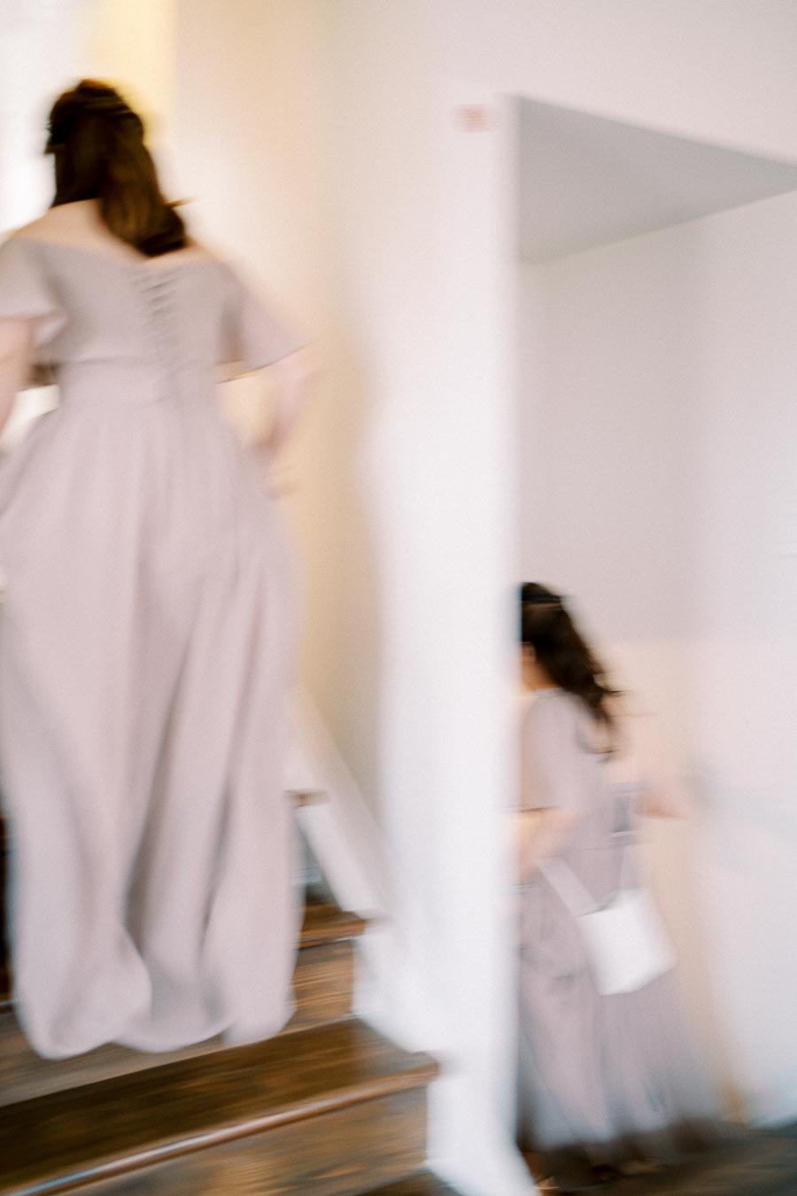 Blurred image of people in light gray dresses ascending wooden stairs in a softly lit interior space, conveying motion and elegance.