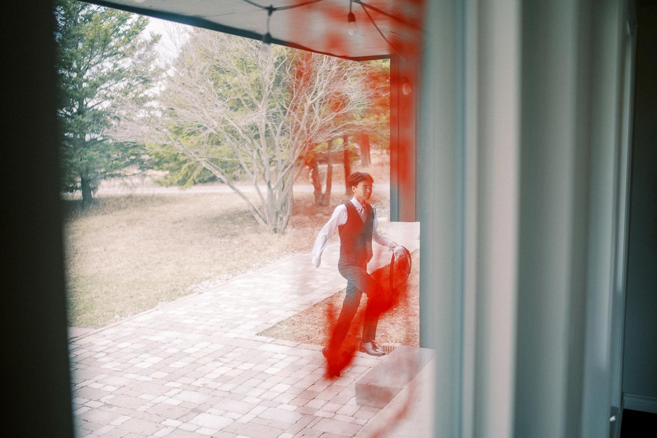 A man stepping onto a patio through a glass door, with blurred red streak along the edge, seen on a reflective window capturing the outdoor scenery of trees and walkway.