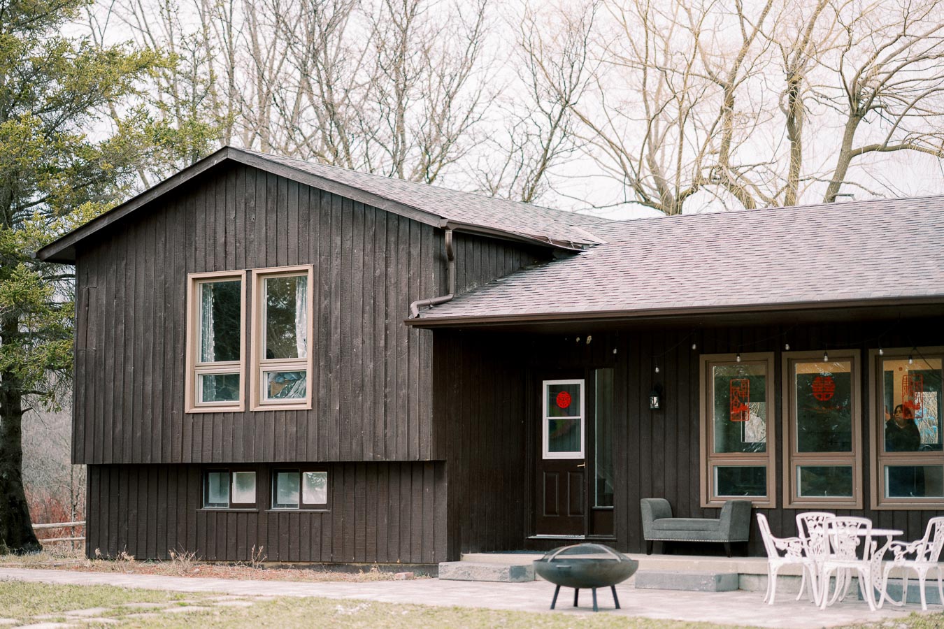 Modern rustic wooden house exterior with large windows and patio furniture in a peaceful wooded setting.