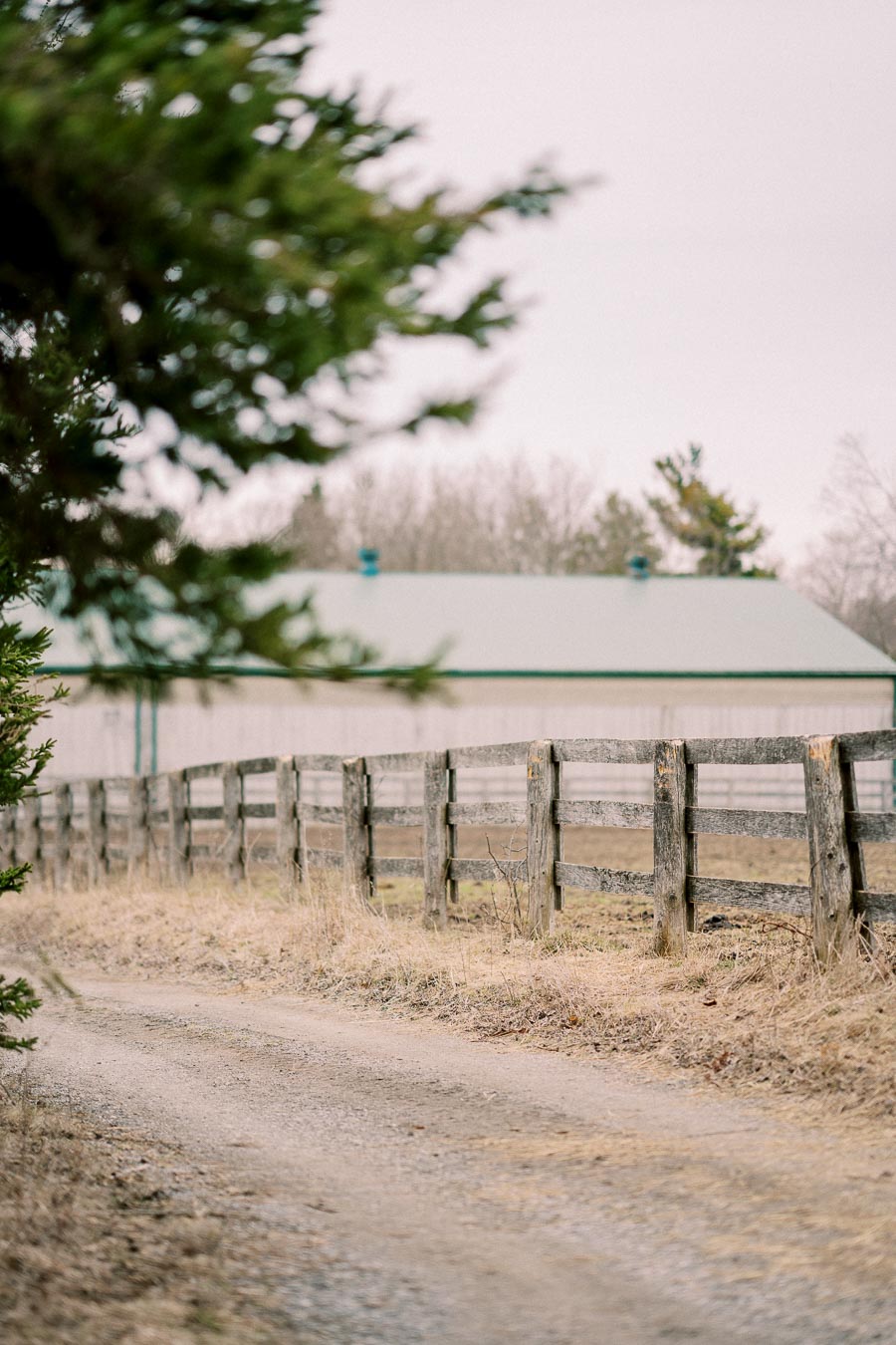 Rustic countryside scene with a wooden fence lining a gravel path leading to a barn, surrounded by trees and dry grass under an overcast sky.