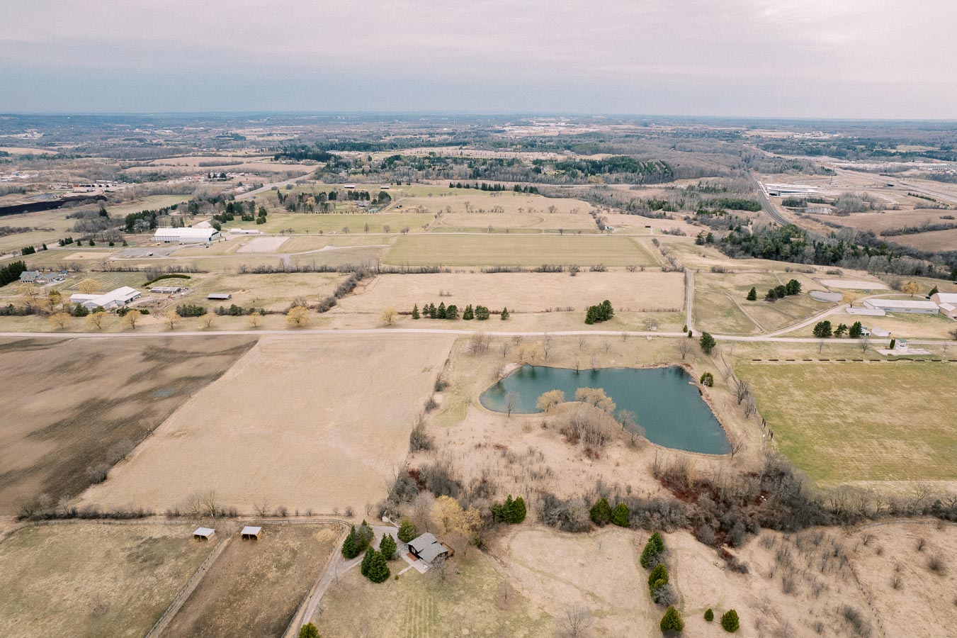 Aerial view of expansive rural landscape featuring green fields, a small pond, scattered trees, and farm buildings under a cloudy sky.