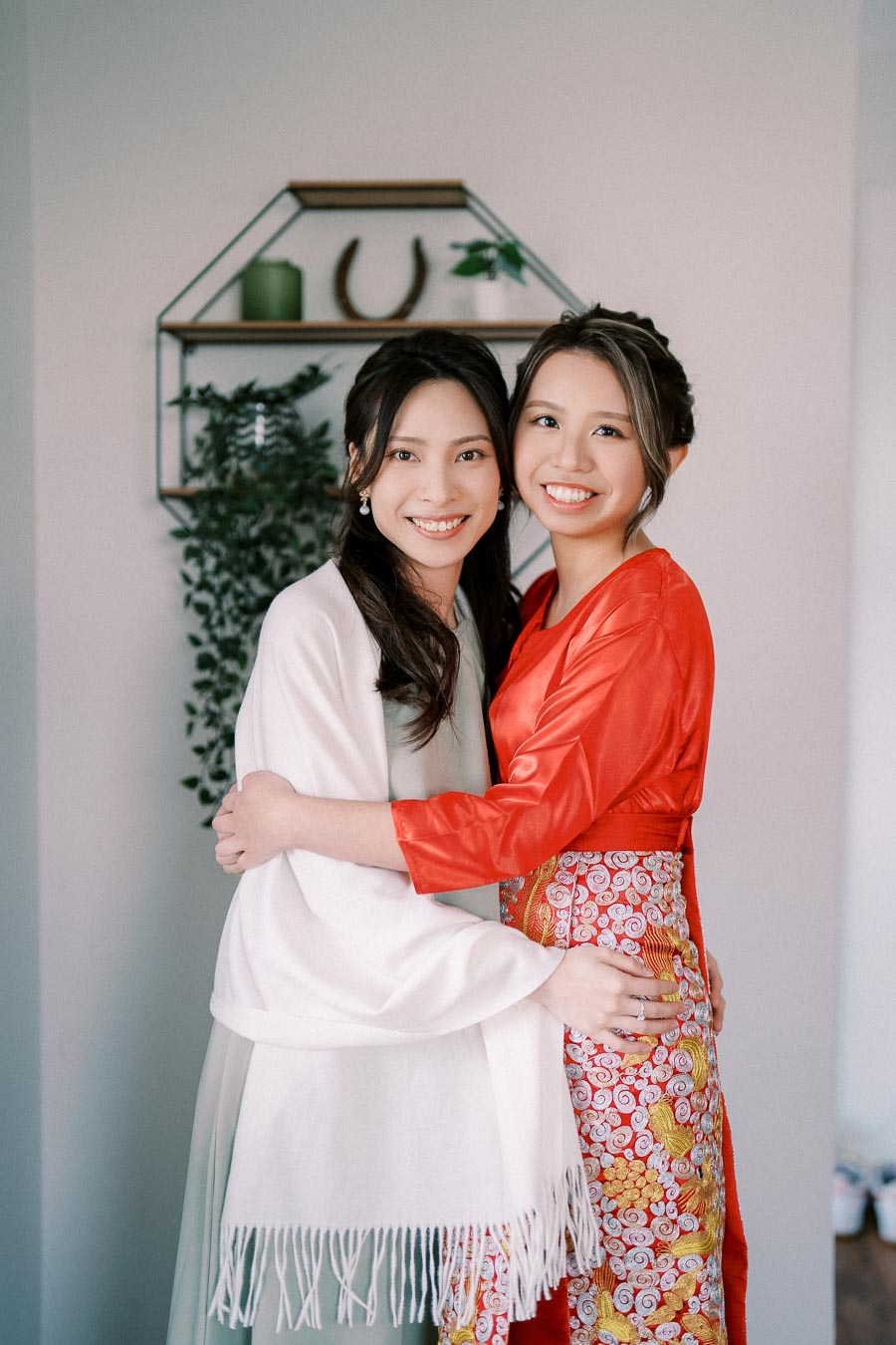 Two women smiling and embracing, one wearing a white shawl and the other in a red traditional dress, standing in a warmly decorated room with a green plant in the background.