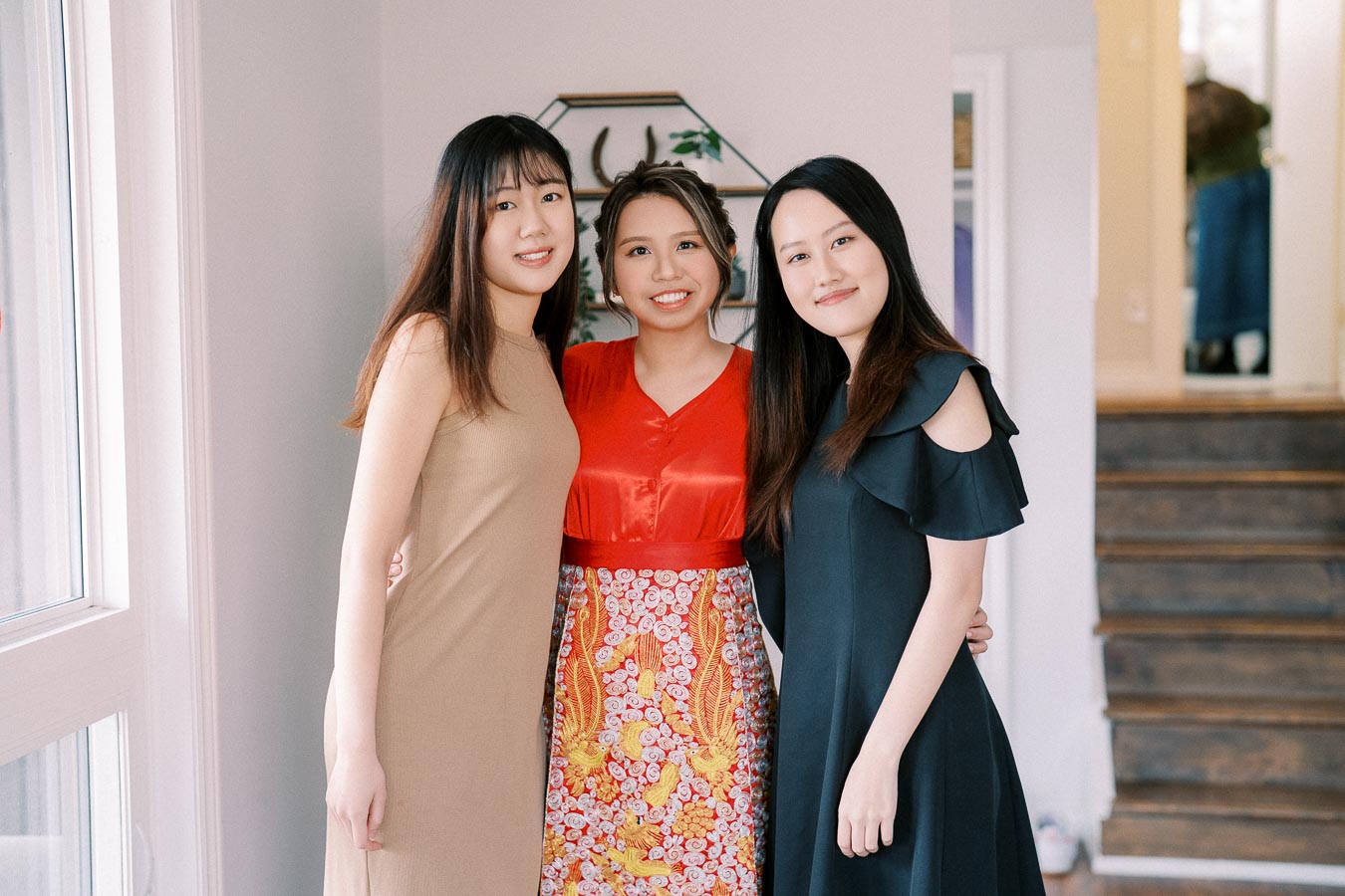 Three young women posing indoors, wearing stylish dresses in beige, red, and black, standing together and smiling in a warmly lit room with wooden stairs in the background.
