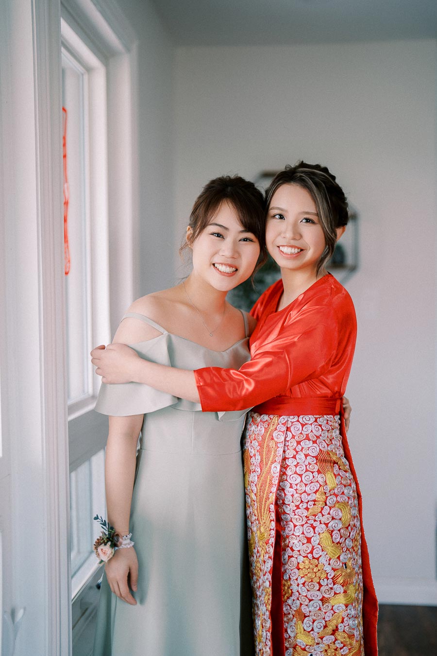 Two smiling women embracing indoors, one wearing a traditional red and gold outfit, and the other in a light grey dress, capturing a joyful moment together.