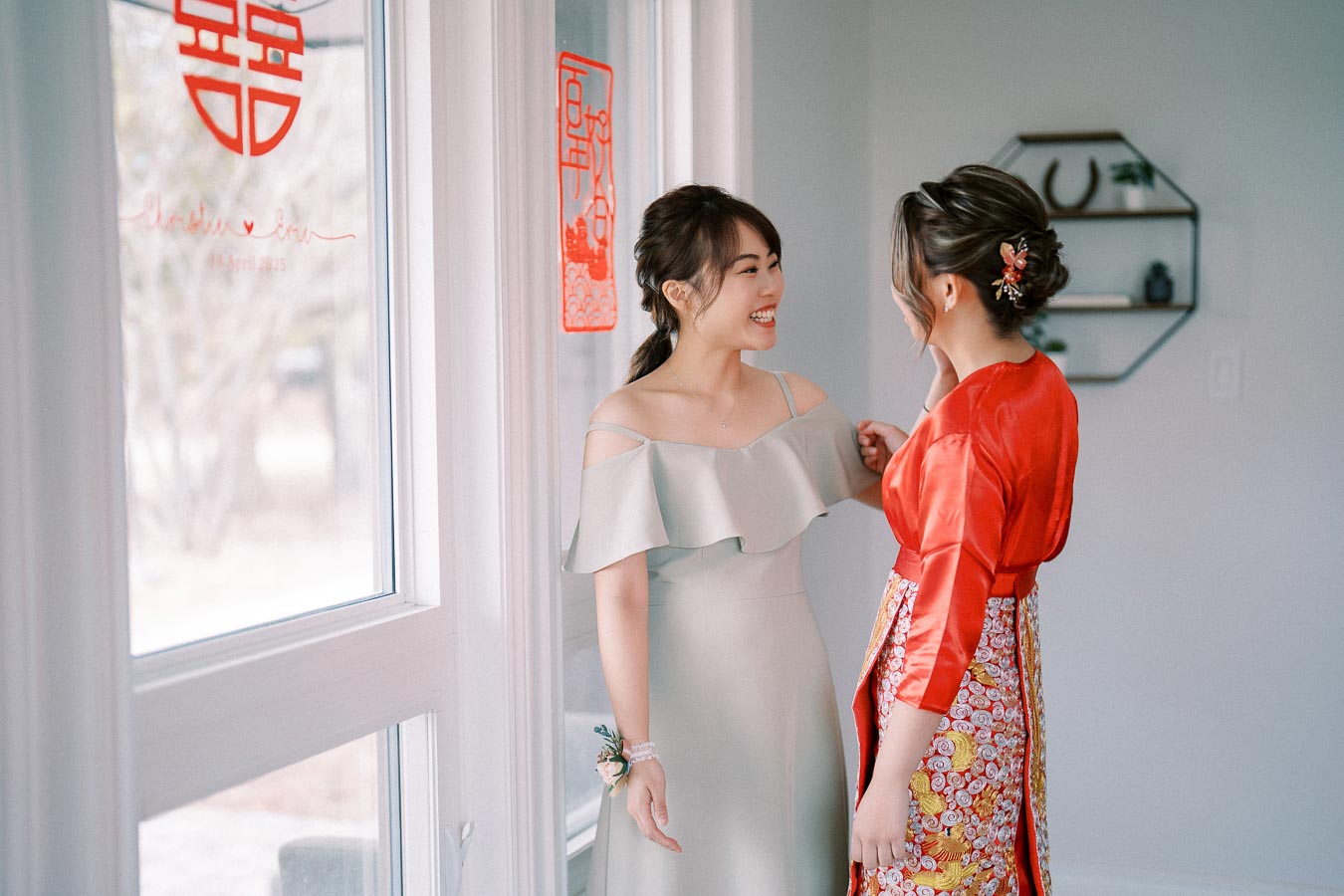 Two women smiling and interacting indoors, one wearing a light grey dress and the other in a traditional red and gold outfit. They stand near a window with Chinese decorations, creating a joyful and festive atmosphere.