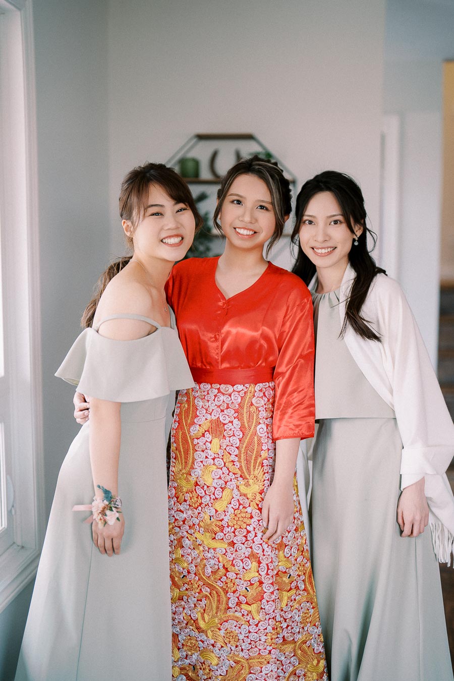 Three smiling women in elegant dresses, one wearing a colorful embroidered outfit with a red top, pose indoors.