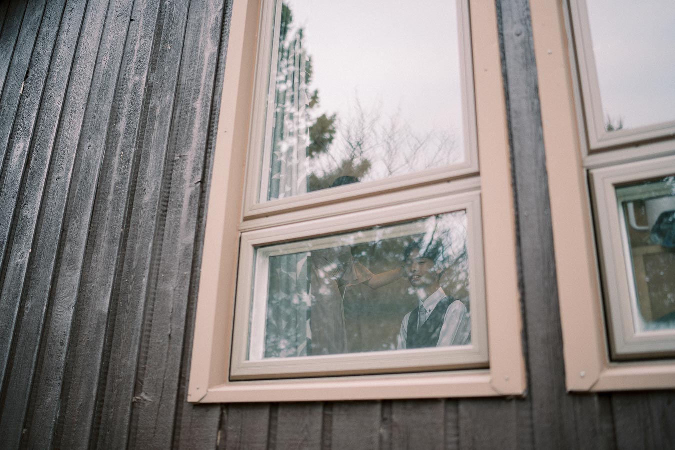 Reflection of a person visible in a window of a rustic wooden building with trees outside, creating a serene and contemplative atmosphere.