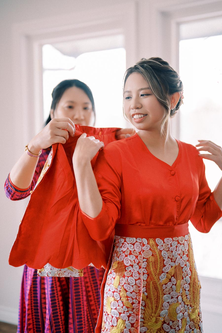 Two women preparing for a traditional event, with one assisting the other in wearing an elaborate red and gold embroidered blouse. The scene captures cultural attire and shared moments in a softly lit room.
