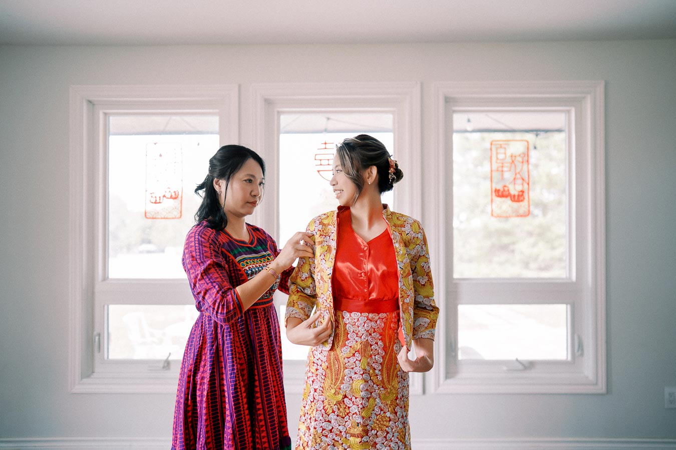 Two women in traditional attire share a moment inside a well-lit room with decorative hangings on the window. One is assisting the other with her colorful outfit, which features intricate patterns. The scene exudes warmth and cultural richness.