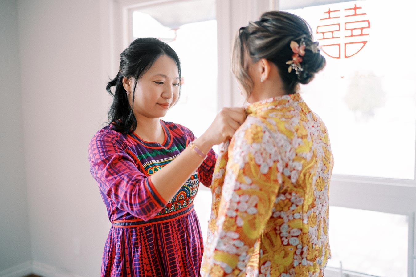 Two women in traditional clothing preparing for a special occasion, with focus on colorful, embroidered attire and a bright room setting. A traditional Chinese symbol is visible in the background, adding cultural significance to the scene.