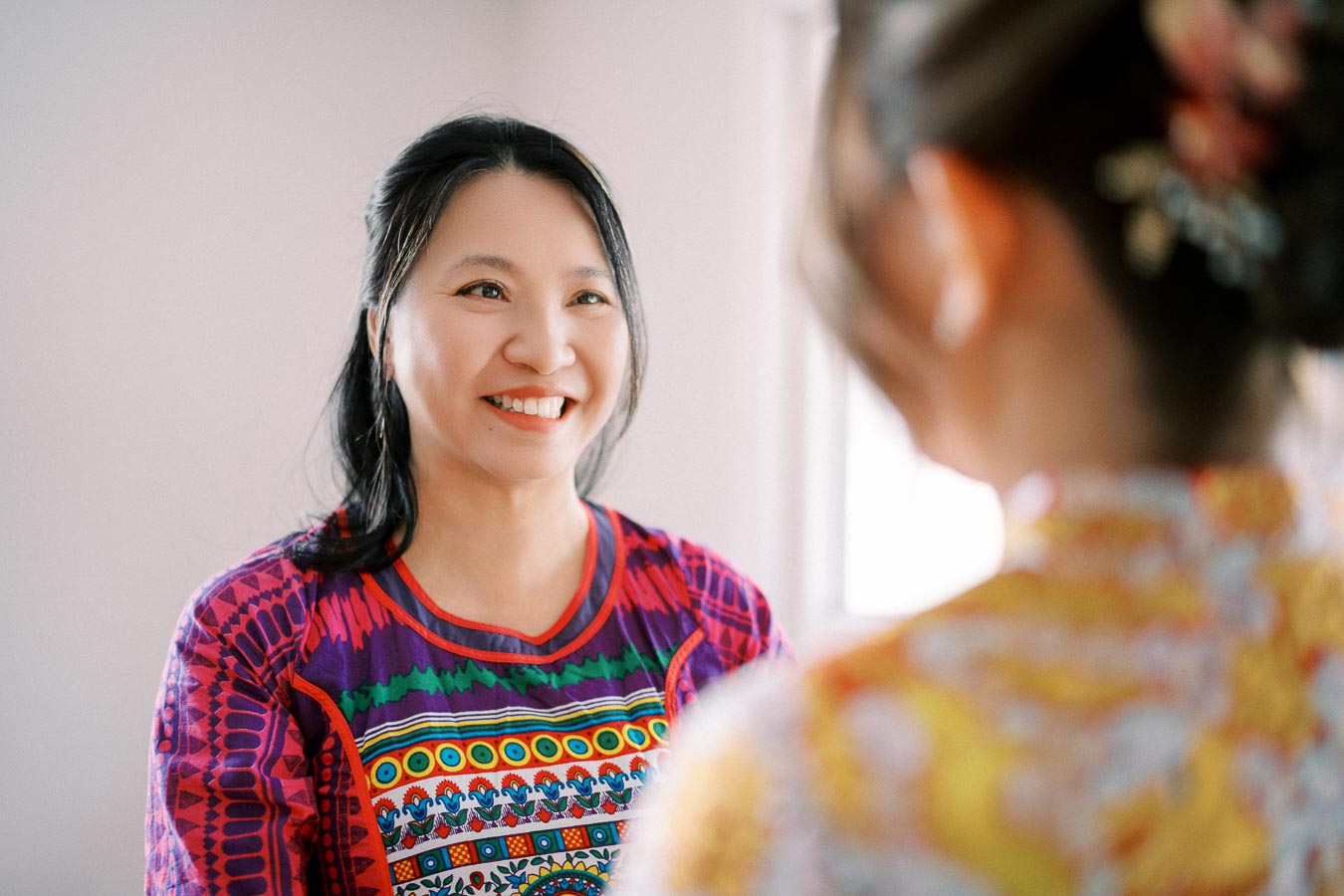 A smiling woman in a colorful patterned shirt looks at another person with a blurred background, creating a joyful and warm atmosphere in the image.