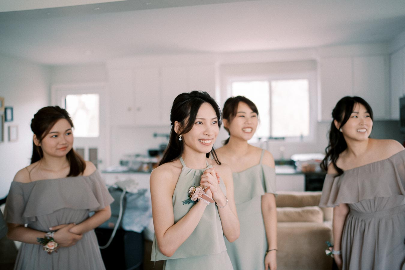 Four smiling women wearing elegant bridesmaid dresses gather in a bright, modern kitchen, showcasing a joyful pre-wedding moment.