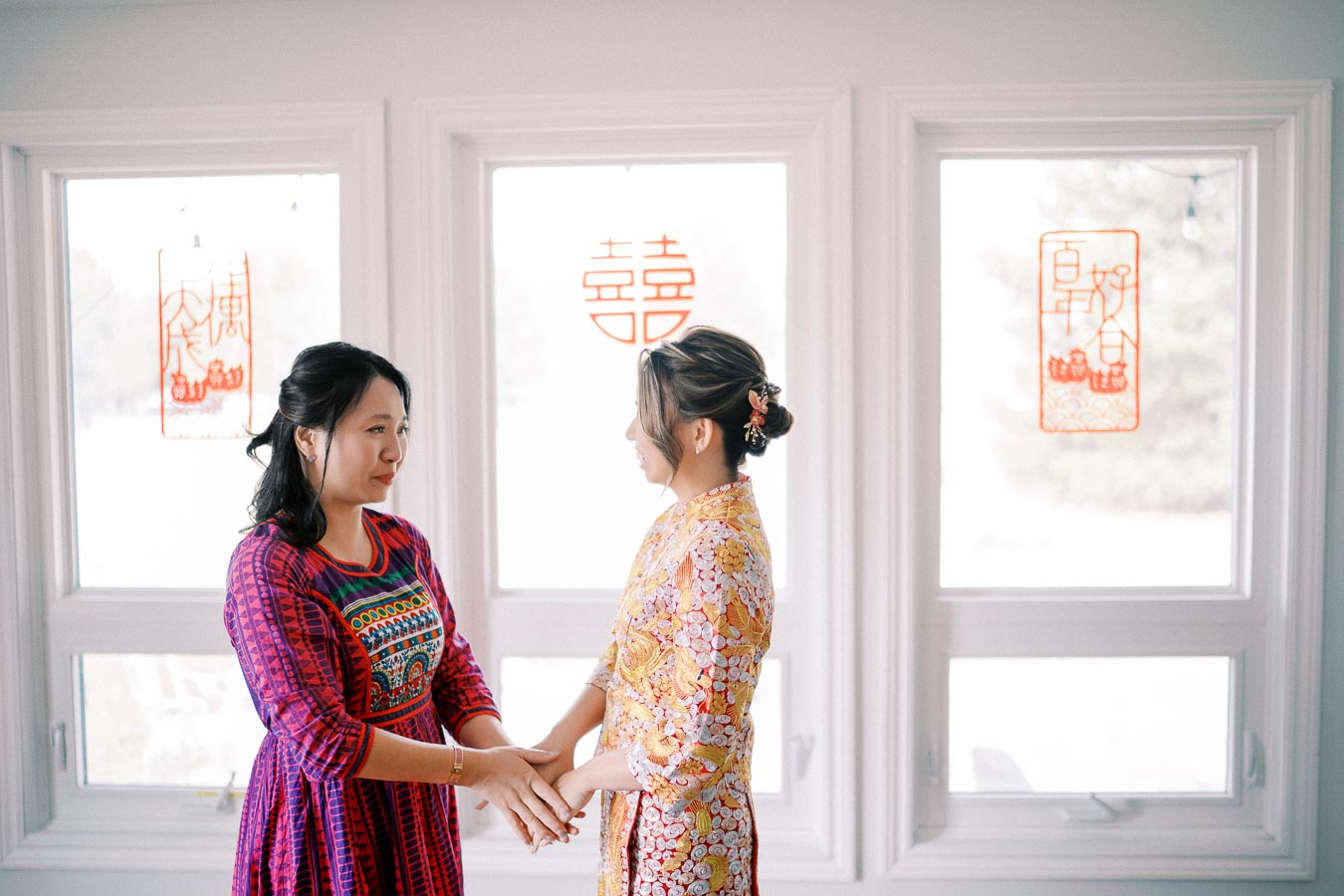 Two women in traditional attire hold hands in a brightly lit room with Chinese decorations on the windows symbolizing happiness.