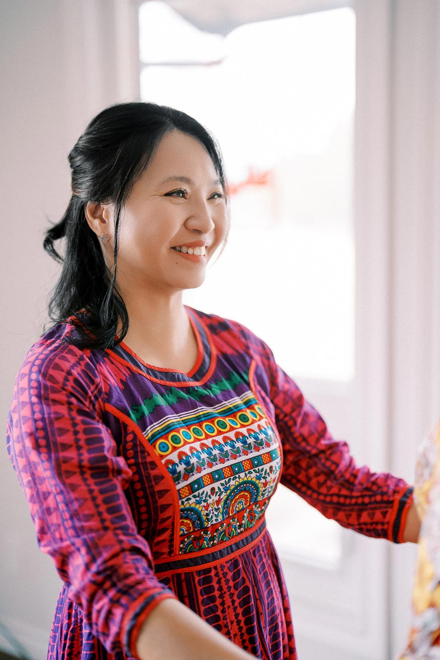 Smiling woman in a colorful, patterned dress with intricate designs standing indoors.
