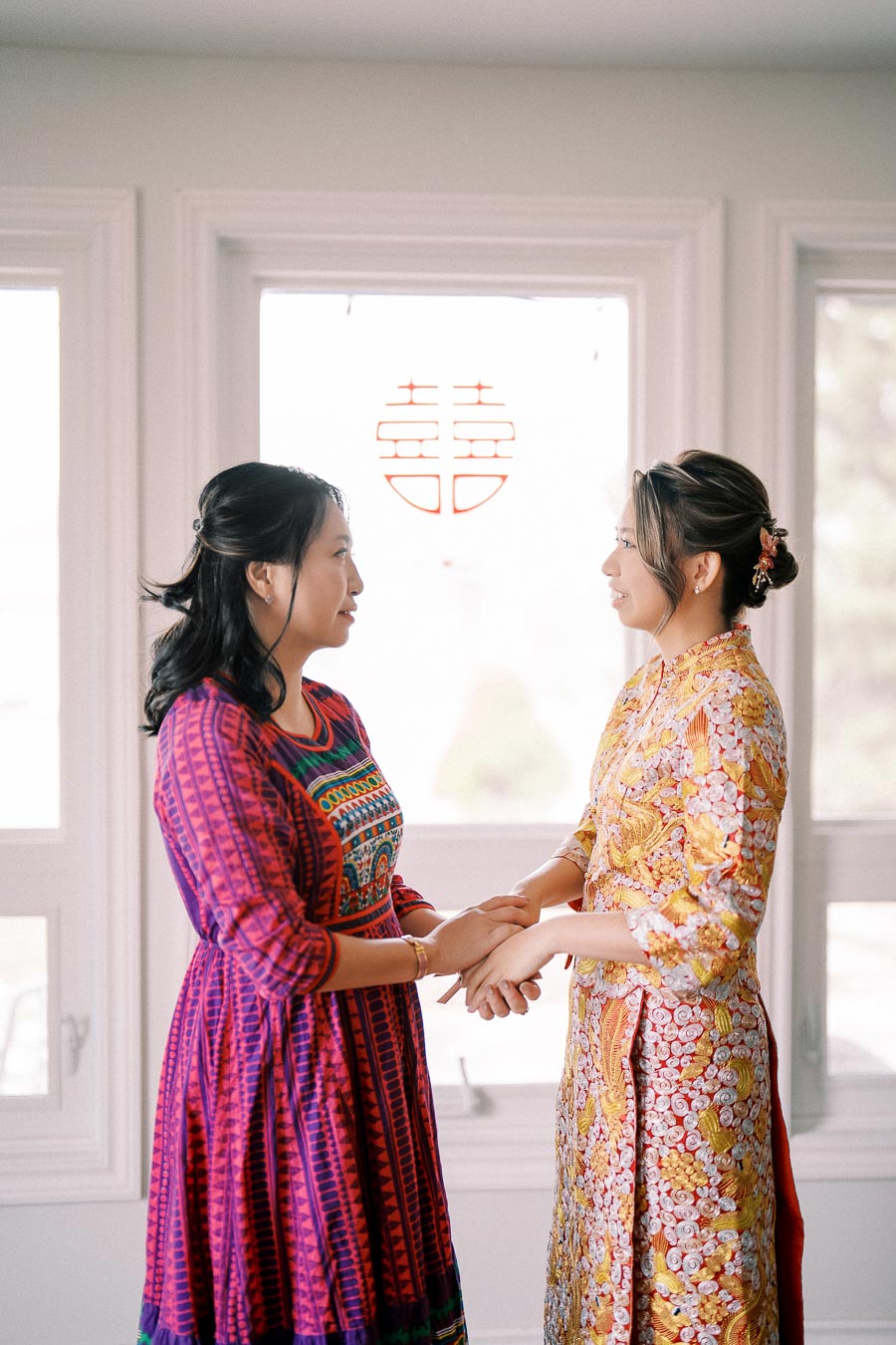 Two women in vibrant traditional dresses share a moment by a window with a double happiness symbol, symbolizing a joyful occasion or cultural celebration.