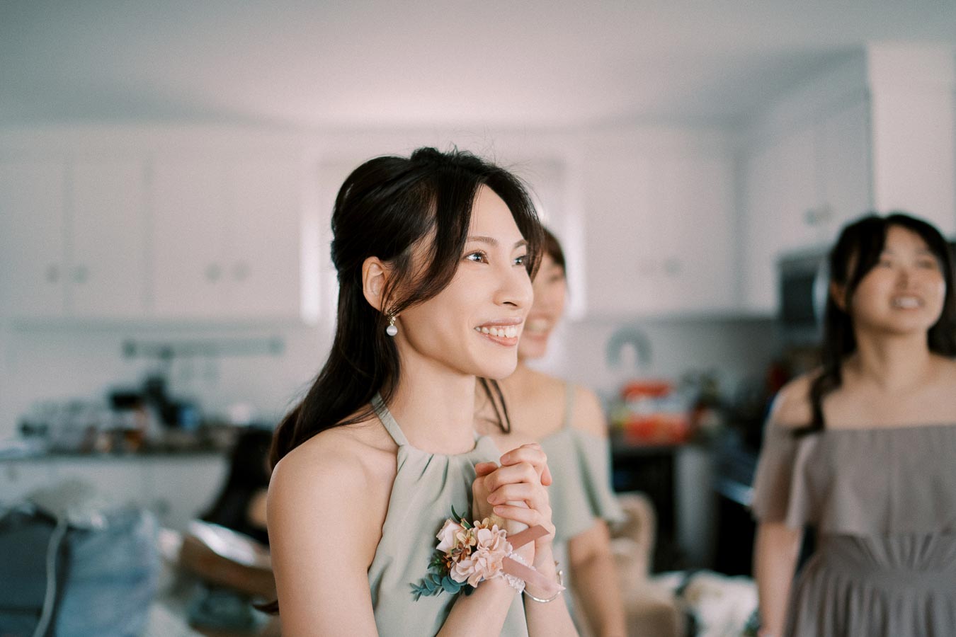 Young woman smiling and looking joyful, wearing a sage green dress with a floral corsage, standing in a bright kitchen with friends in the background.