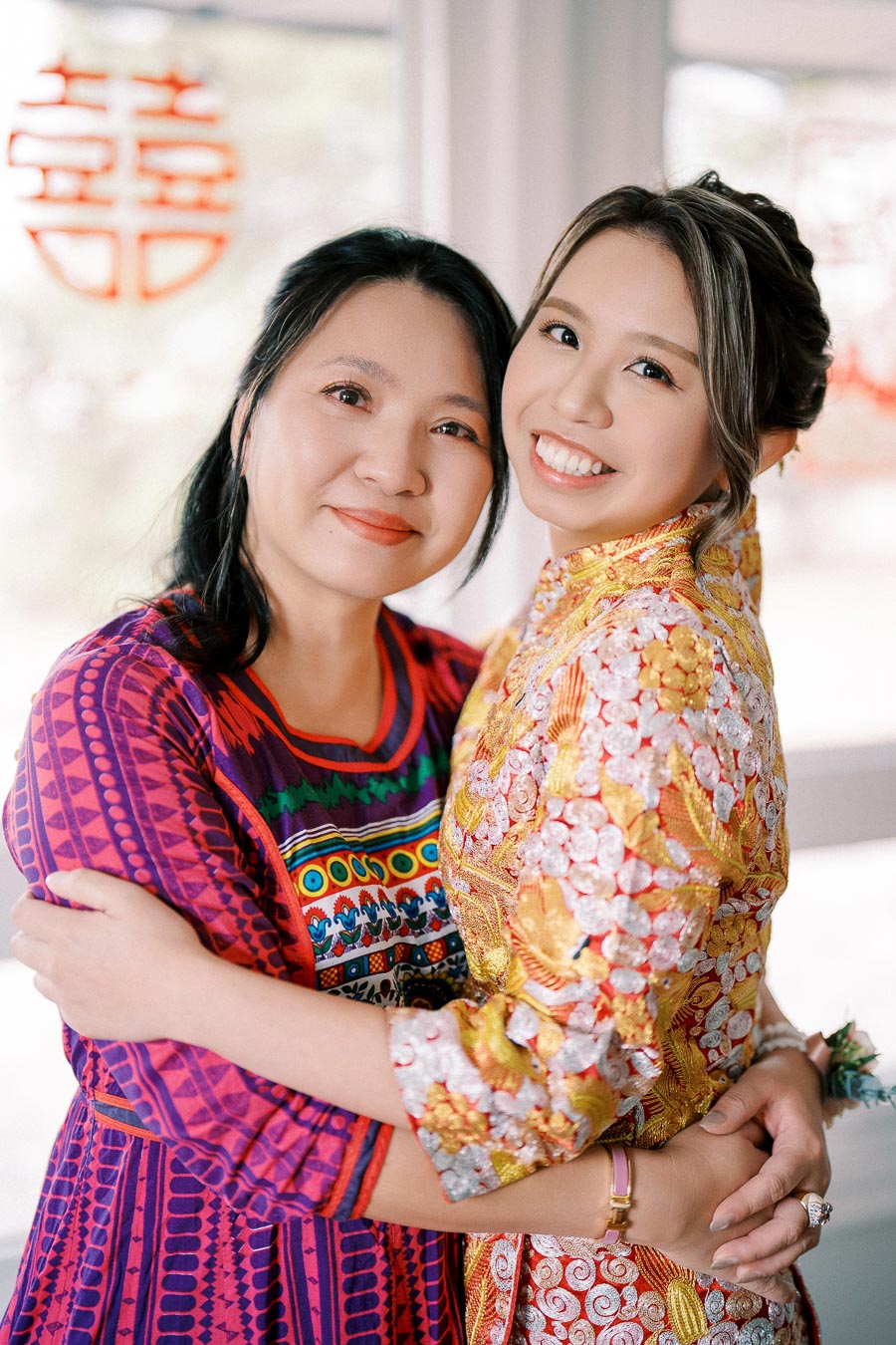 A smiling bride in a traditional golden wedding dress embraces a woman in a colorful patterned outfit, symbolizing love and cultural celebration.