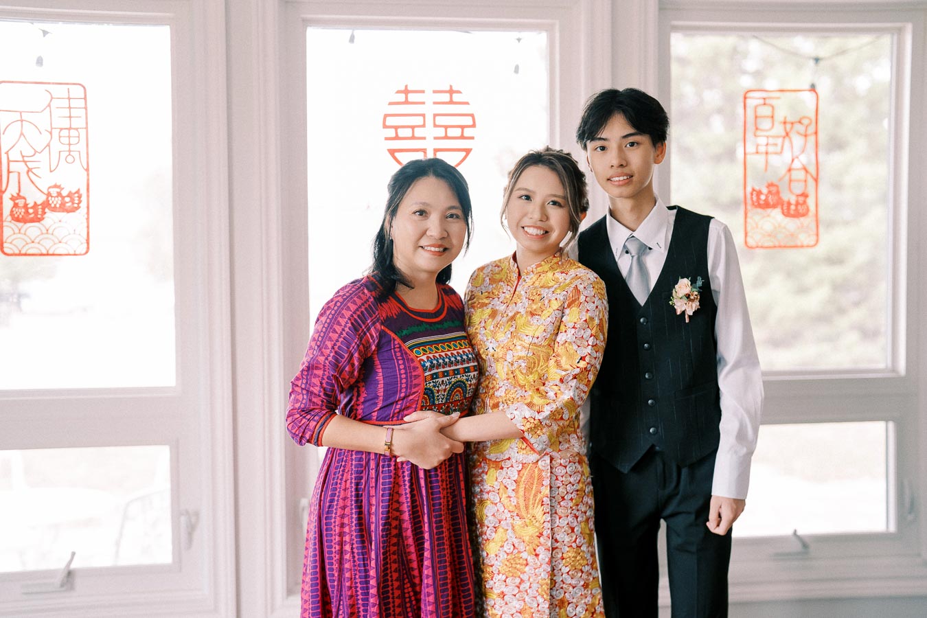 A group photo featuring three people standing together, dressed in vibrant traditional attire. The background shows Chinese symbols on the windows, possibly indicating a celebration or cultural event. One person is in a colorful dress, another in an elegant embroidered yellow outfit, and the third wearing a suit. They are smiling, conveying happiness and togetherness.