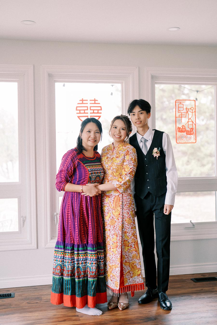 A family standing together indoors, with a woman in a colorful patterned dress, a bride in a traditional floral embroidered outfit, and a young man in a suit, set against a backdrop of bright windows with festive red Chinese decorations.