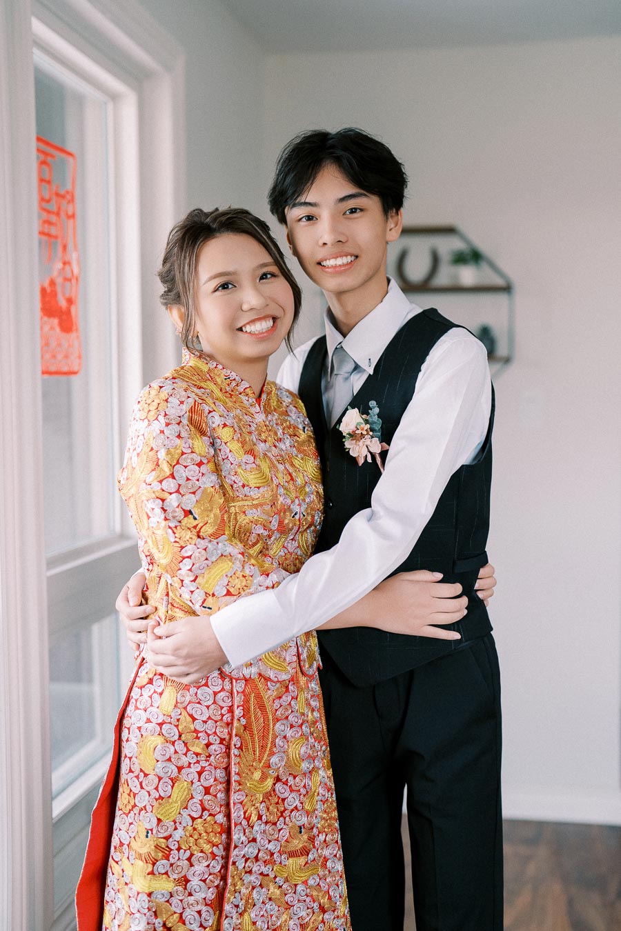 Young couple in traditional Vietnamese attire smiling and embracing in a bright room, with festive decorations visible in the background.