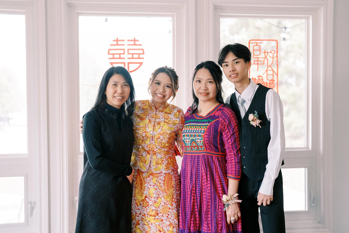 A group of four people posing in colorful traditional attire during a celebration, with Chinese symbols decorating the windows in the background.