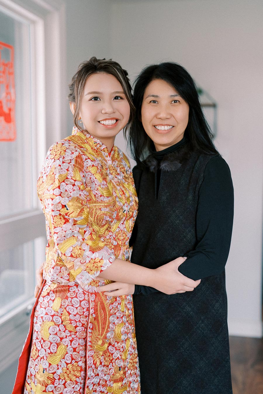 Two women smiling and embracing each other; one wearing a traditional Chinese dress in red and gold embroidery, the other in a black outfit, standing in a brightly lit room.