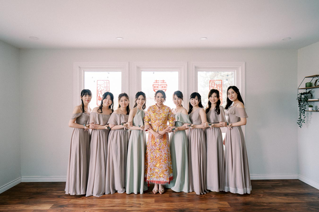 A bride in a traditional Chinese dress standing with her bridesmaids in matching taupe dresses, posing together indoors with Chinese characters on the windows in the background, celebrating a wedding.