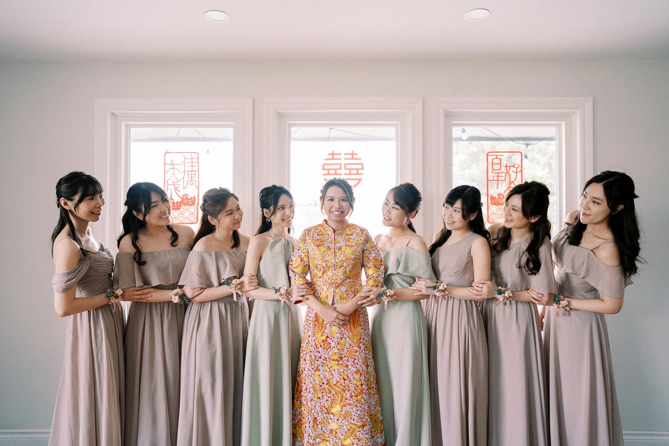 Bride in traditional yellow and pink floral dress surrounded by bridesmaids in lavender dresses, standing indoors with decorative red Chinese characters on windows, smiling happily.