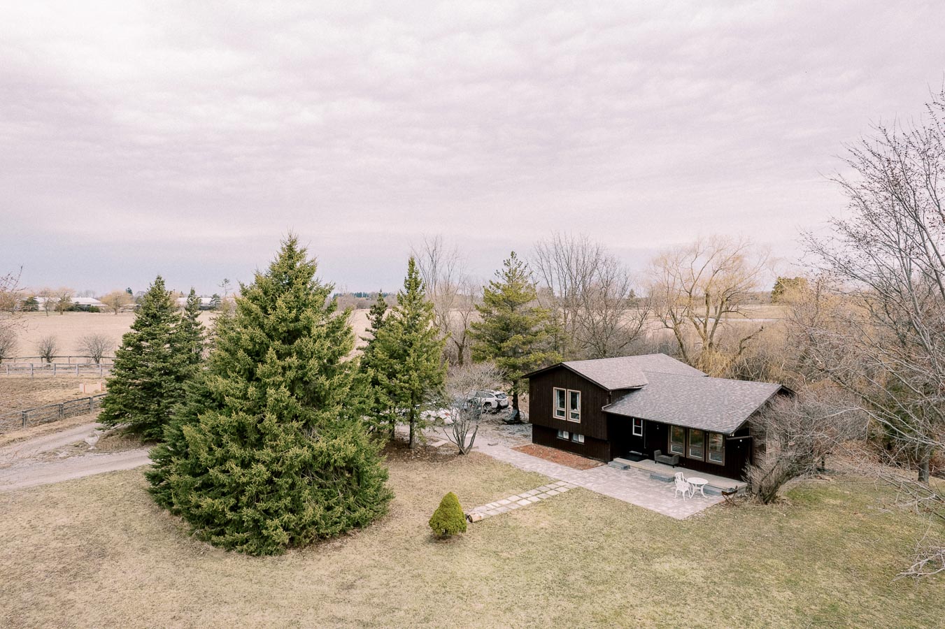 Aerial view of a cozy rural cabin surrounded by lush green trees and expansive open fields under a cloudy sky.