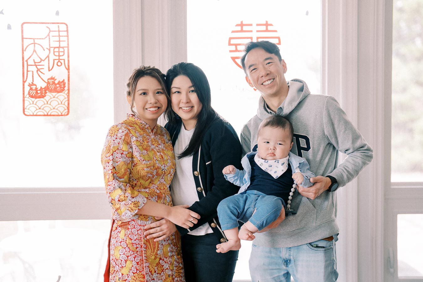A happy family posing together indoors with traditional Asian decorations, including a woman in a floral dress and a man holding a baby, with bright window light in the background.