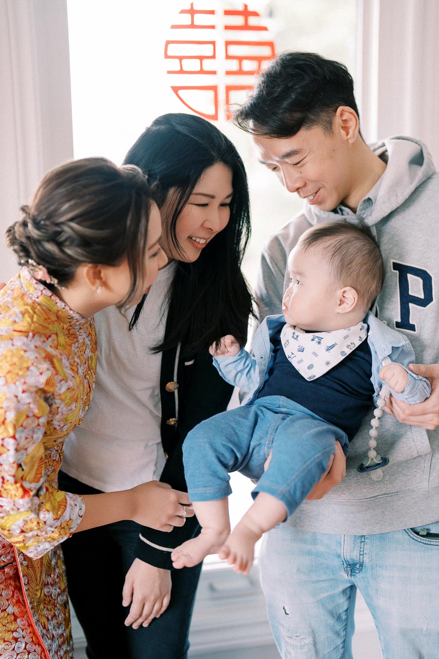 A family gathering around a baby dressed in blue, with parents and relatives smiling warmly indoors, creating a joyful and loving atmosphere.