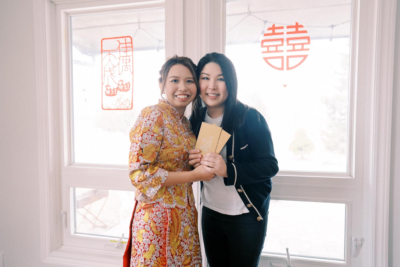 Two women smiling and holding cards in a festive setting, with traditional Chinese decorations on the window. One is wearing a colorful, patterned outfit, suggesting a celebration or cultural event.