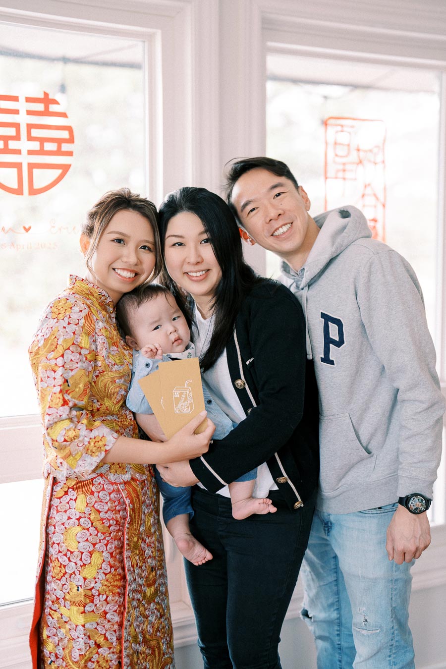 A happy family celebrating a special occasion, with a woman in a traditional embroidered dress holding a baby next to two people standing by her side, in front of a window with decorative red symbols.
