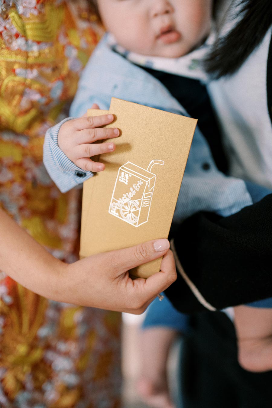 A child holding a golden envelope with a white design, assisted by an adult wearing ornate traditional attire, possibly during a cultural celebration or festive occasion.