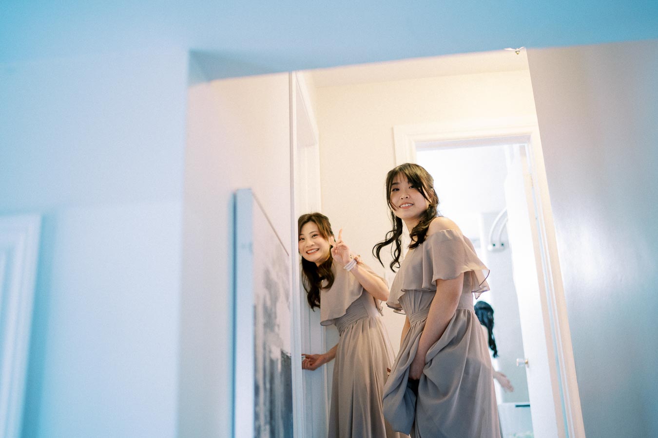 Two women in elegant dresses smile as they pose in a brightly lit doorway, with one making a peace sign, creating a cheerful and relaxed atmosphere.