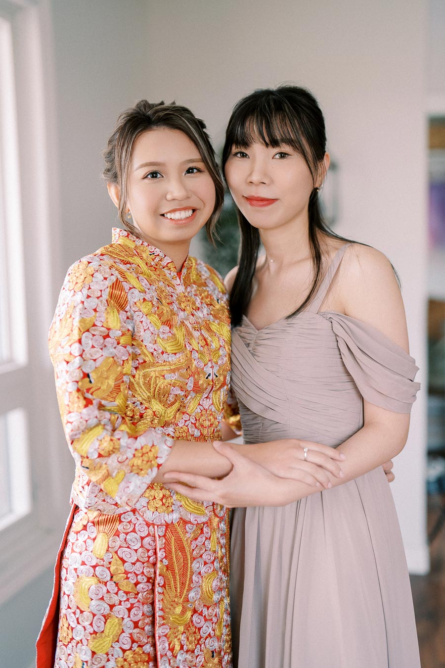 Two women smiling and embracing; one in a traditional colorful embroidered dress, and the other in a light, elegant gown, indoors near a window.
