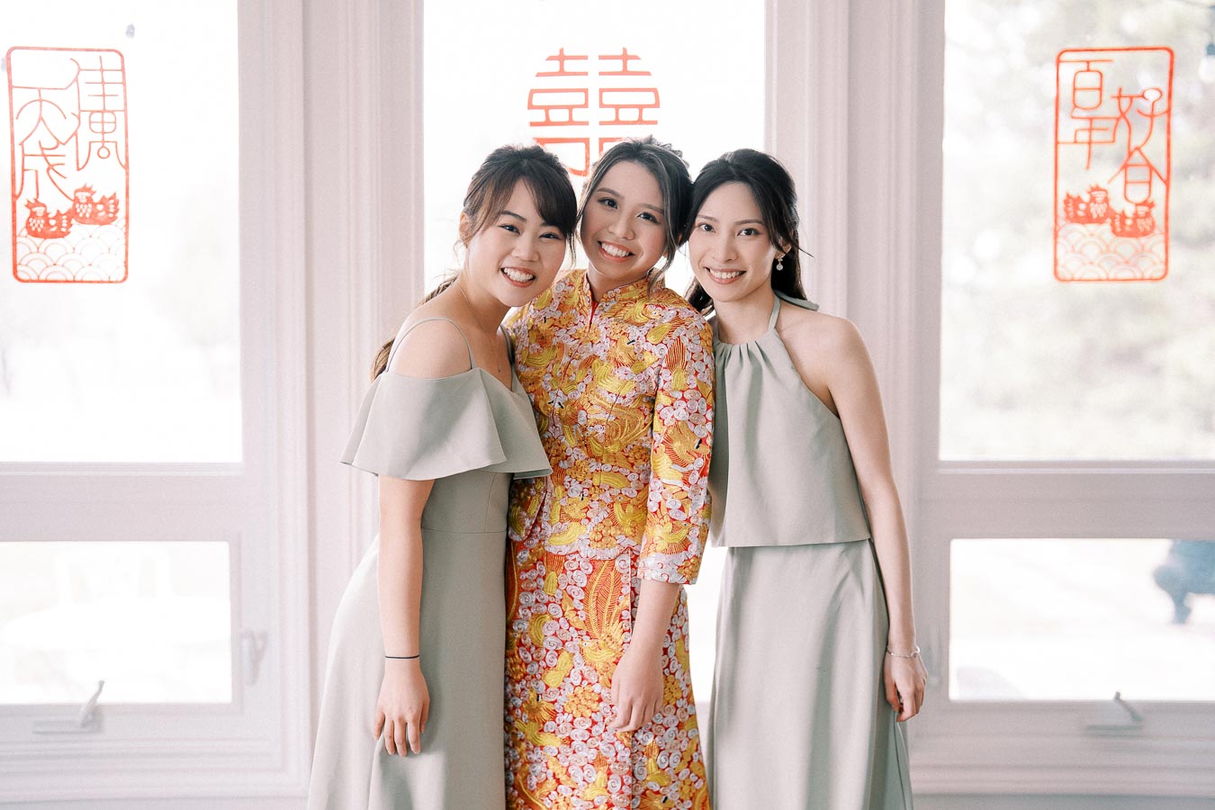 Three women in elegant dresses pose together in front of a window with festive red Chinese symbols, capturing a joyous occasion. The central woman wears a colorful traditional dress, suggesting a celebration or wedding. The setting features soft lighting and a harmonious atmosphere.