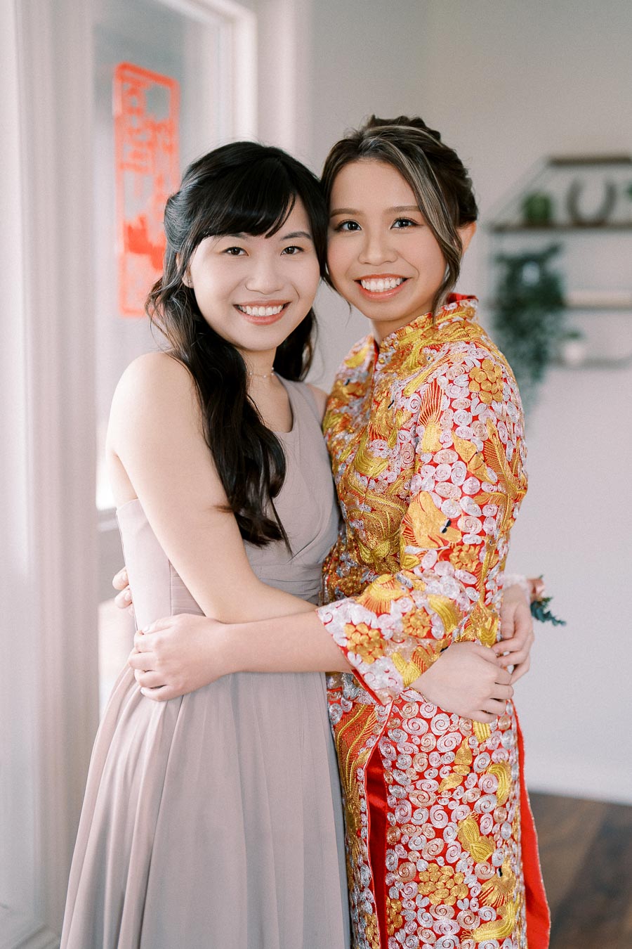Two women smiling and embracing, one in a light gown and the other in a traditional colorful embroidered dress, standing in a well-lit room with minimal decor.