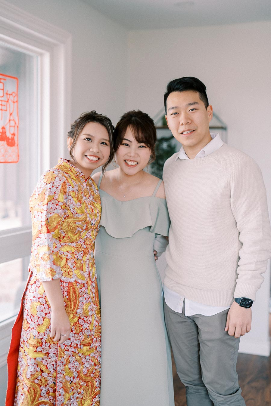 Three people smiling indoors, with one woman wearing a colorful traditional dress, another woman in a light green dress, and a man in a light sweater. The image depicts a happy moment during a likely special event or celebration.
