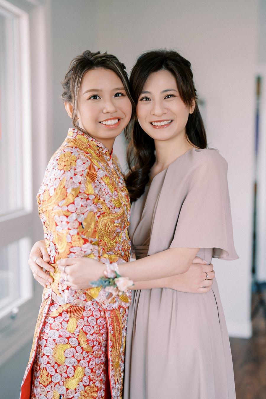 Two women smiling and embracing indoors, one wearing an elegant traditional embroidered dress and the other in a light-colored modern outfit, under soft natural lighting.