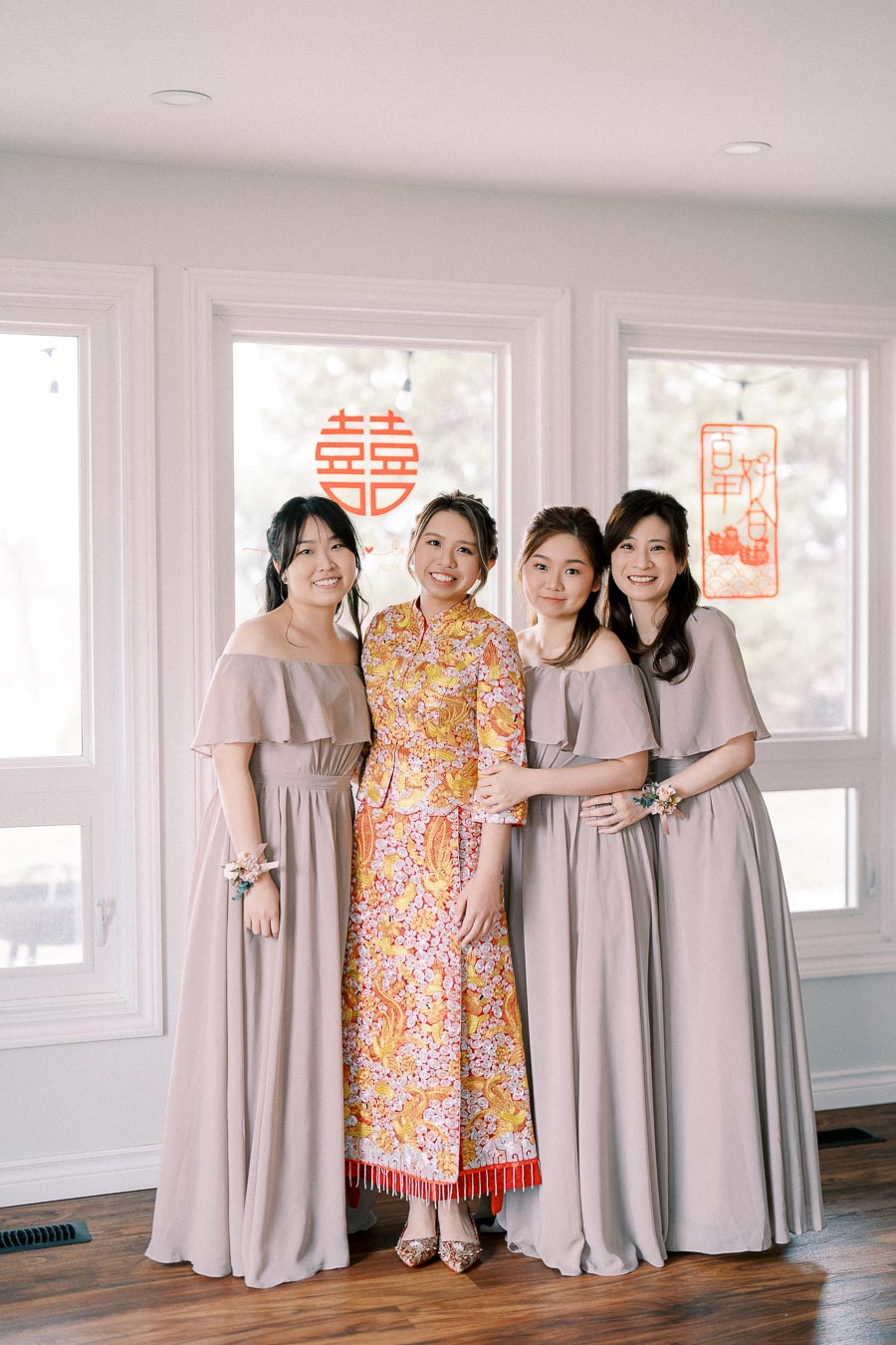 Four women posing together indoors at a traditional wedding, with the bride wearing a colorful floral dress and bridesmaids dressed in matching taupe gowns. Chinese characters for double happiness are displayed on the window behind them, symbolizing celebration and joy.