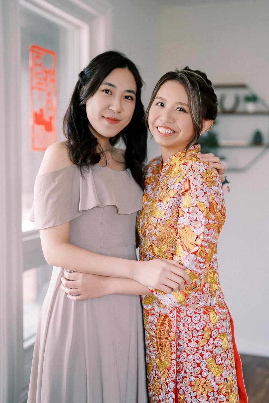 Two women smiling and embracing; one is wearing a lavender off-shoulder dress and the other is in a colorful traditional dress with intricate patterns, standing in a bright indoor setting.