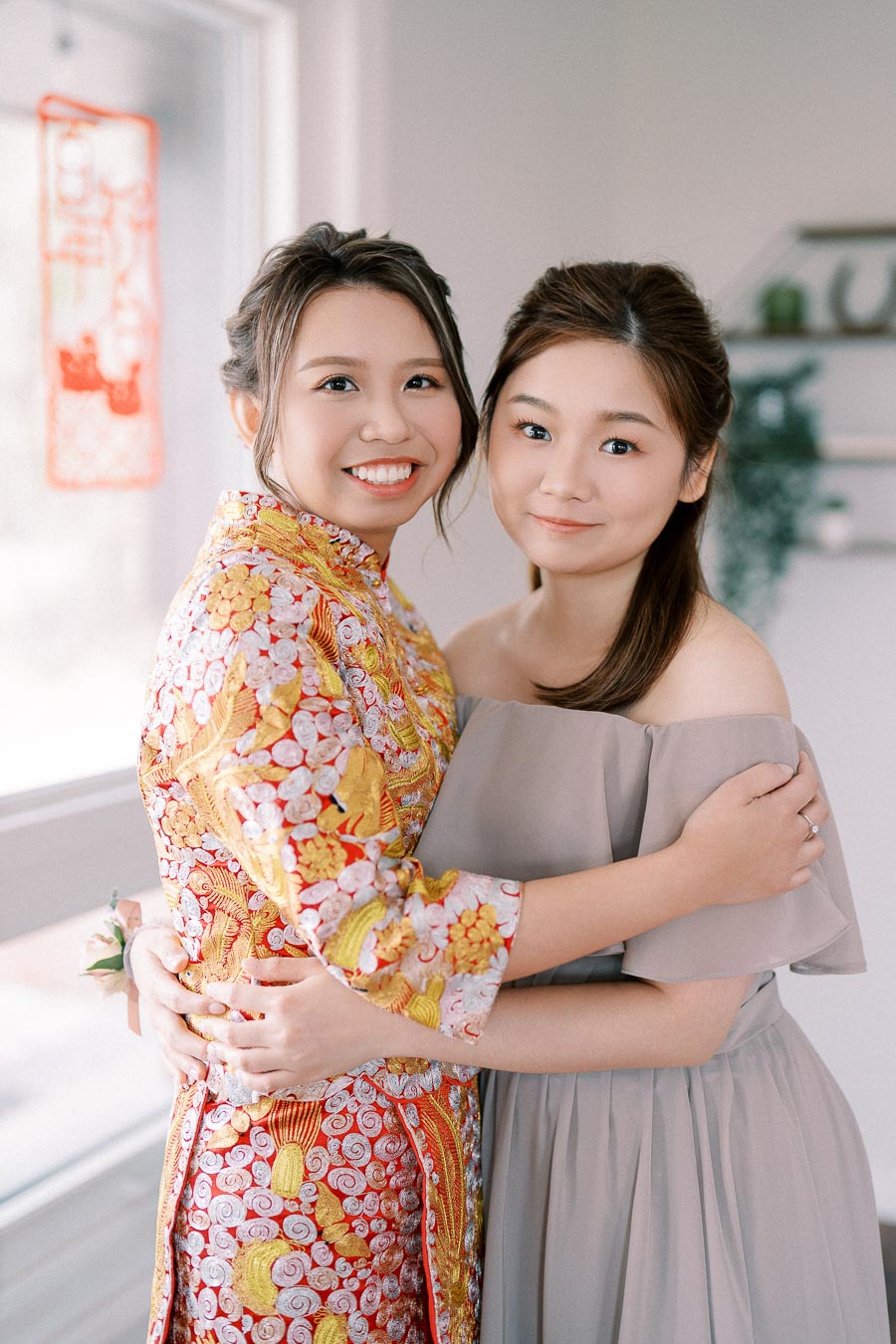 Two smiling women embracing, one wearing a traditional colorful dress with intricate gold and red embroidery, and the other in a light gray off-shoulder gown, standing indoors with soft natural light.