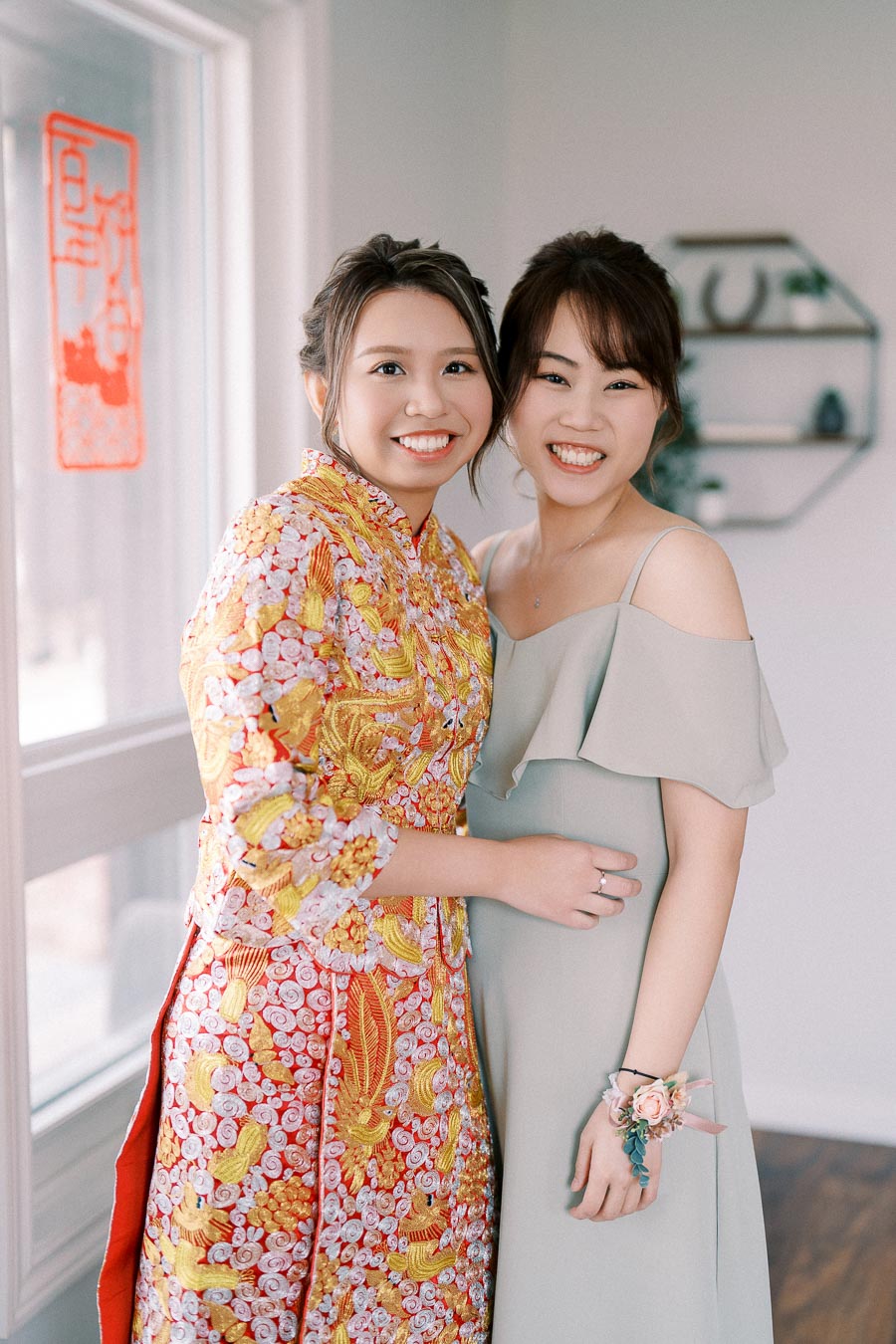 Two people smiling, wearing elegant dresses, one in a traditional embroidered outfit and the other in a modern light-colored gown, standing together indoors next to a window with decorative elements in the background.