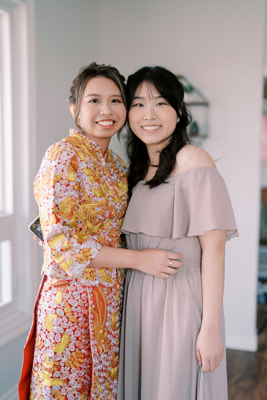 Two women smiling and embracing; one wears a traditional Vietnamese dress with colorful embroidery while the other is in a light gray off-shoulder gown, standing indoors.