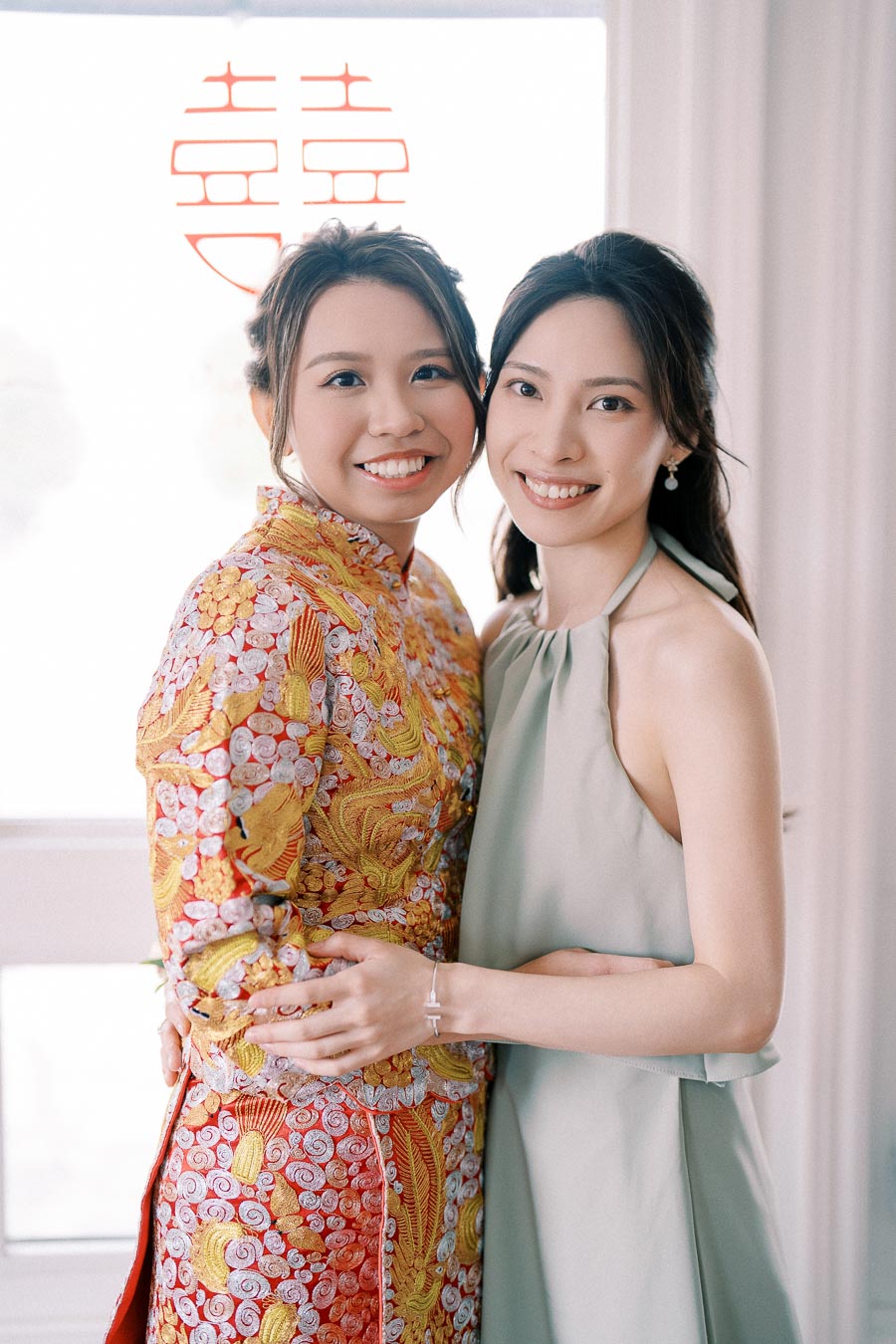 Two smiling women embracing, one wearing a traditional red and gold embroidered dress, symbolizing a cultural celebration, with a Chinese double happiness symbol in the background.