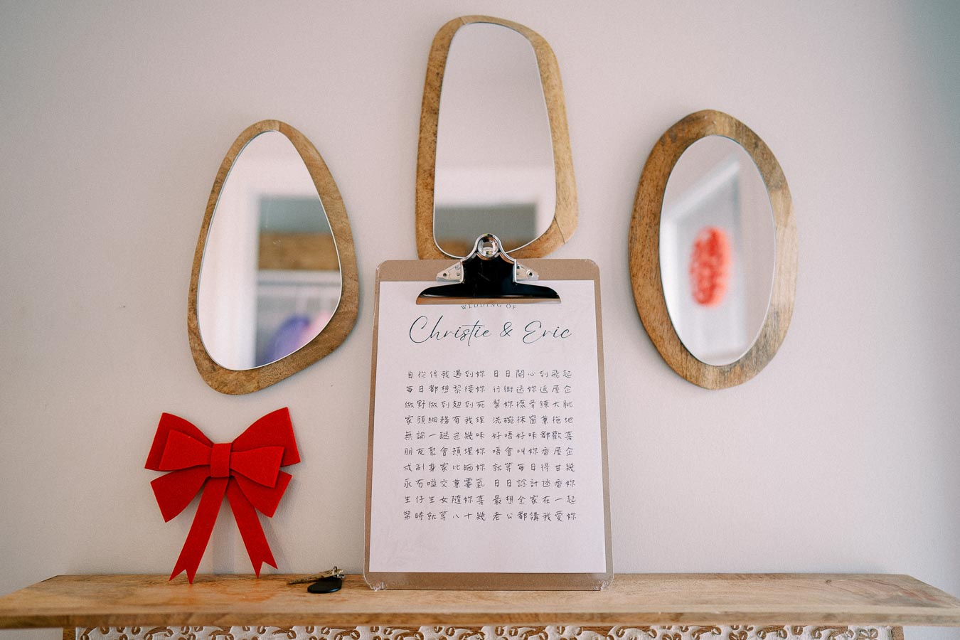 A decorative display featuring three wooden-framed mirrors with a clipboard in the center. The clipboard holds a paper titled Wedding of Christie & Eric with text in Chinese script. A bright red ribbon bow is placed on a wooden shelf below. Modern home decor for a special event.