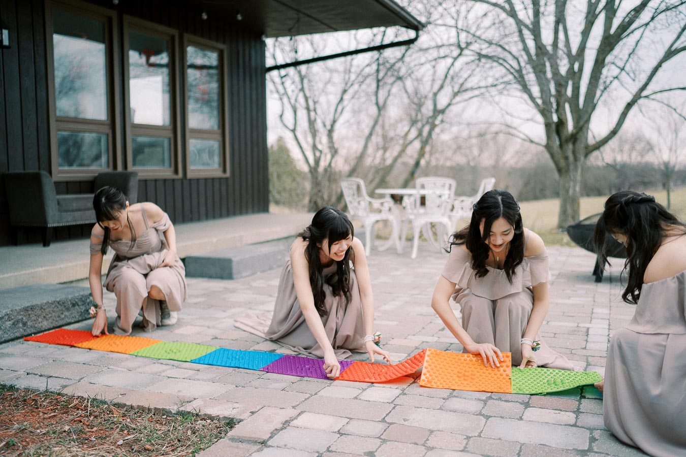 Four women in matching beige dresses arrange colorful interlocking foam mats on an outdoor patio, with a cozy cabin and trees in the background.