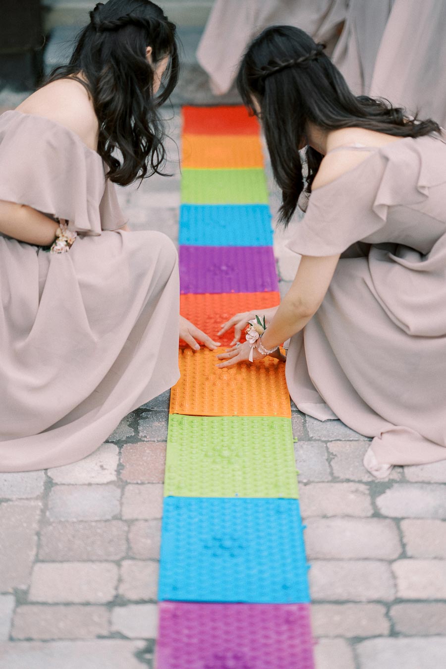 Two women in formal dresses arranging colorful, interlocking mats on a cobblestone path.